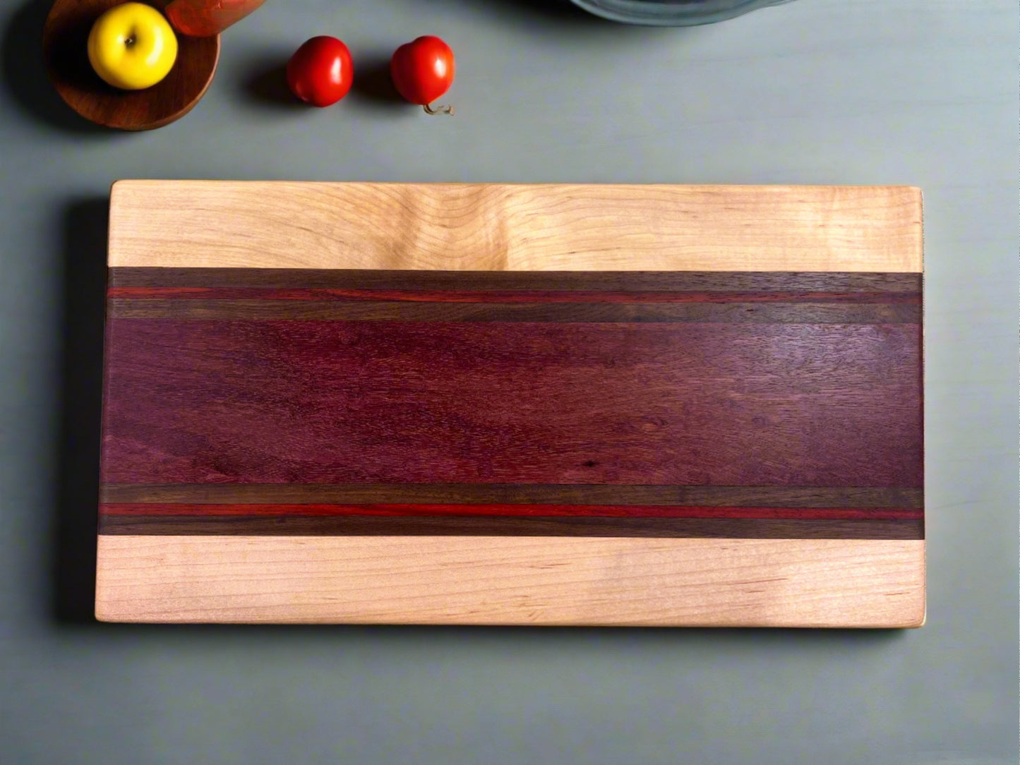 Artisan Board #2 by Wet Nose Woodworks, featuring a bold dark wood stripe, sits on a gray surface with two red tomatoes and a yellow fruit in a dish at the top left—showcasing vibrant colors and fine craftsmanship.