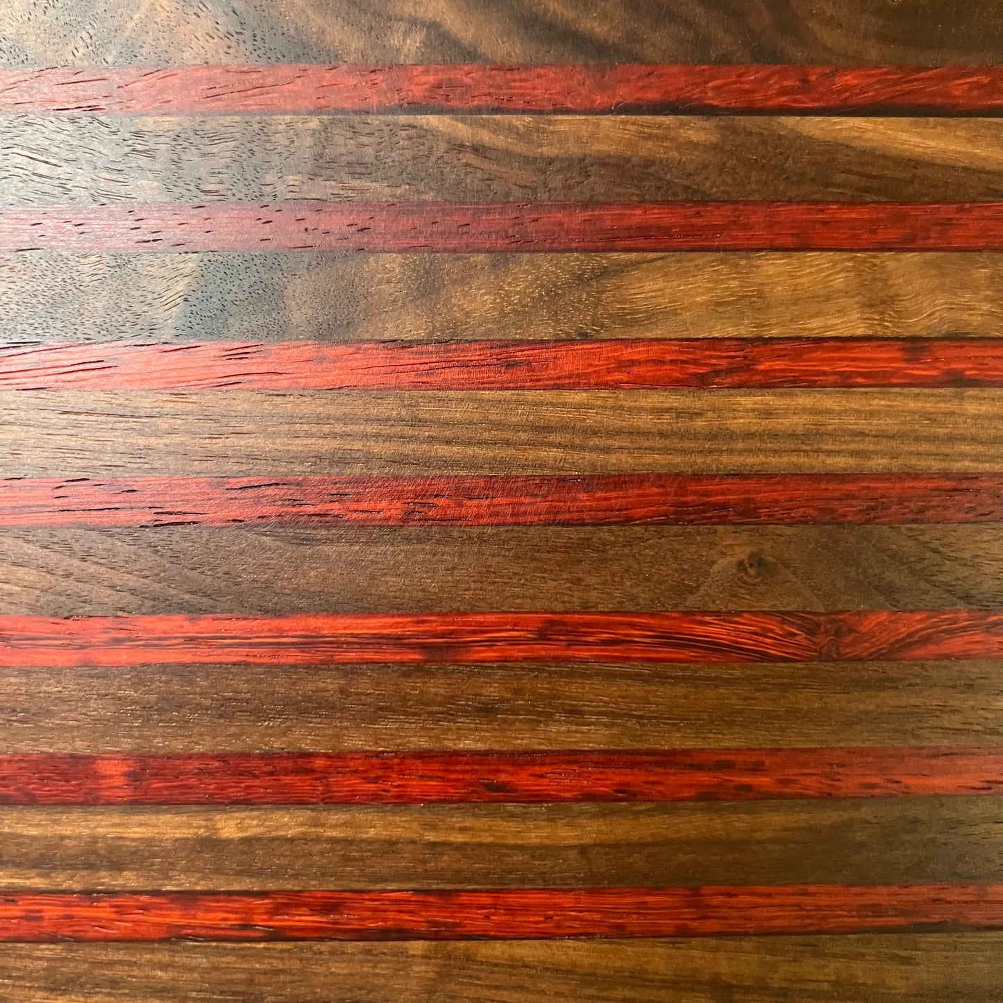 The Wet Nose Woodworks Artisan Board #7, a rectangular cheese board with a handle and hanging hole, features alternating walnut and padauk stripes and is placed on a light marble surface.