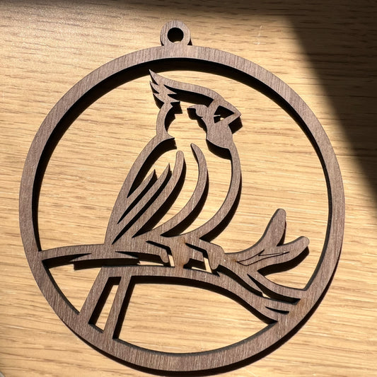 The Wet Nose Woodworks Cardinal ornament is a round, black handcrafted wooden piece featuring a cardinal silhouette on a branch, perfect for hanging in your window against an outdoor backdrop.