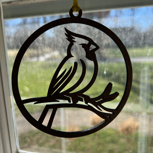 The Wet Nose Woodworks Cardinal ornament is a round, black handcrafted wooden piece featuring a cardinal silhouette on a branch, perfect for hanging in your window against an outdoor backdrop.