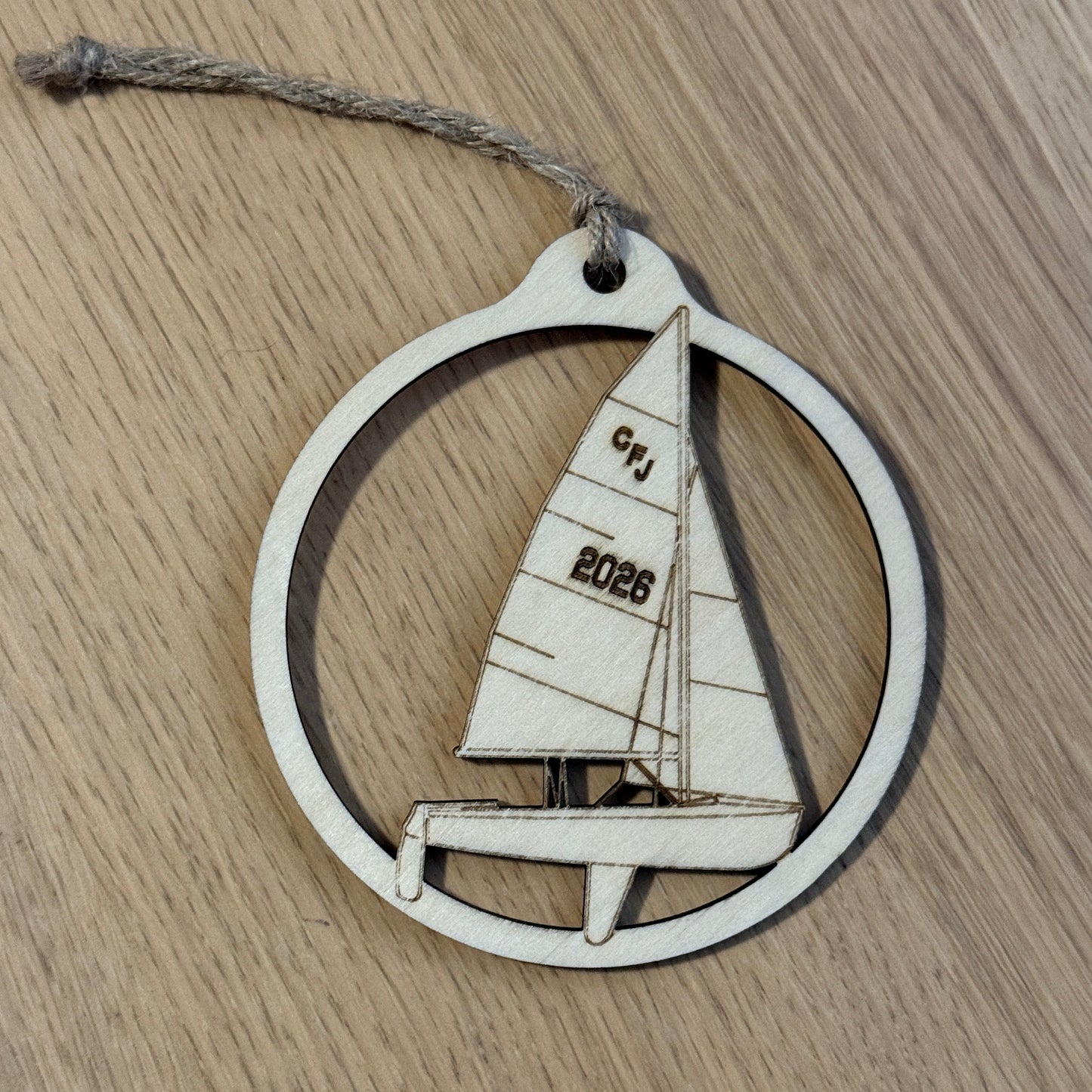 A circular wooden ornament featuring a laser-cut sailboat design with the engraved text 'CFJ' and '2026', hanging from a rustic twine string on a light wood grain surface.