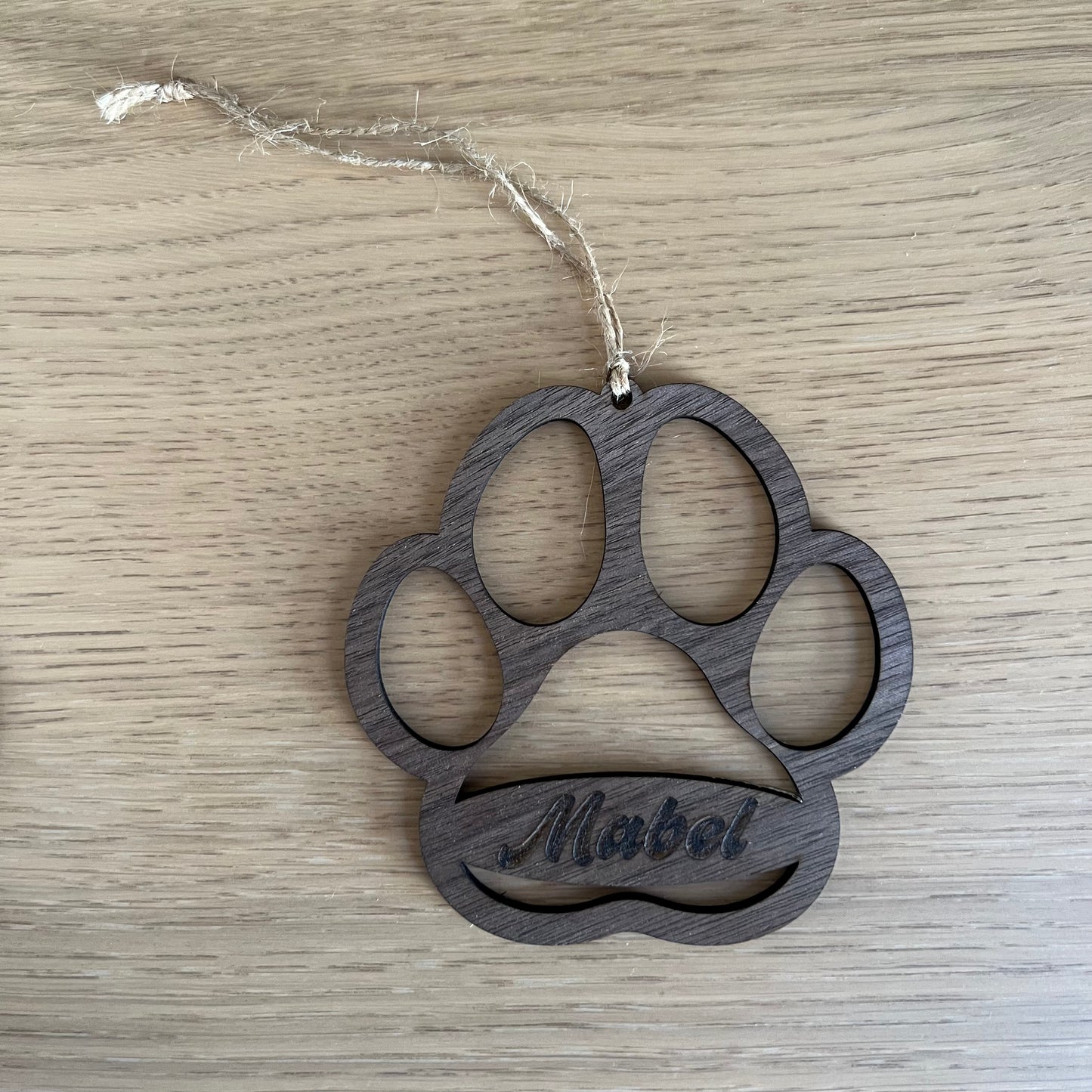 A hand hangs a personalized Dog Paw ornament by Wet Nose Woodworks with the name Mabel on a green Christmas tree, adding a unique touch to your holiday decor.