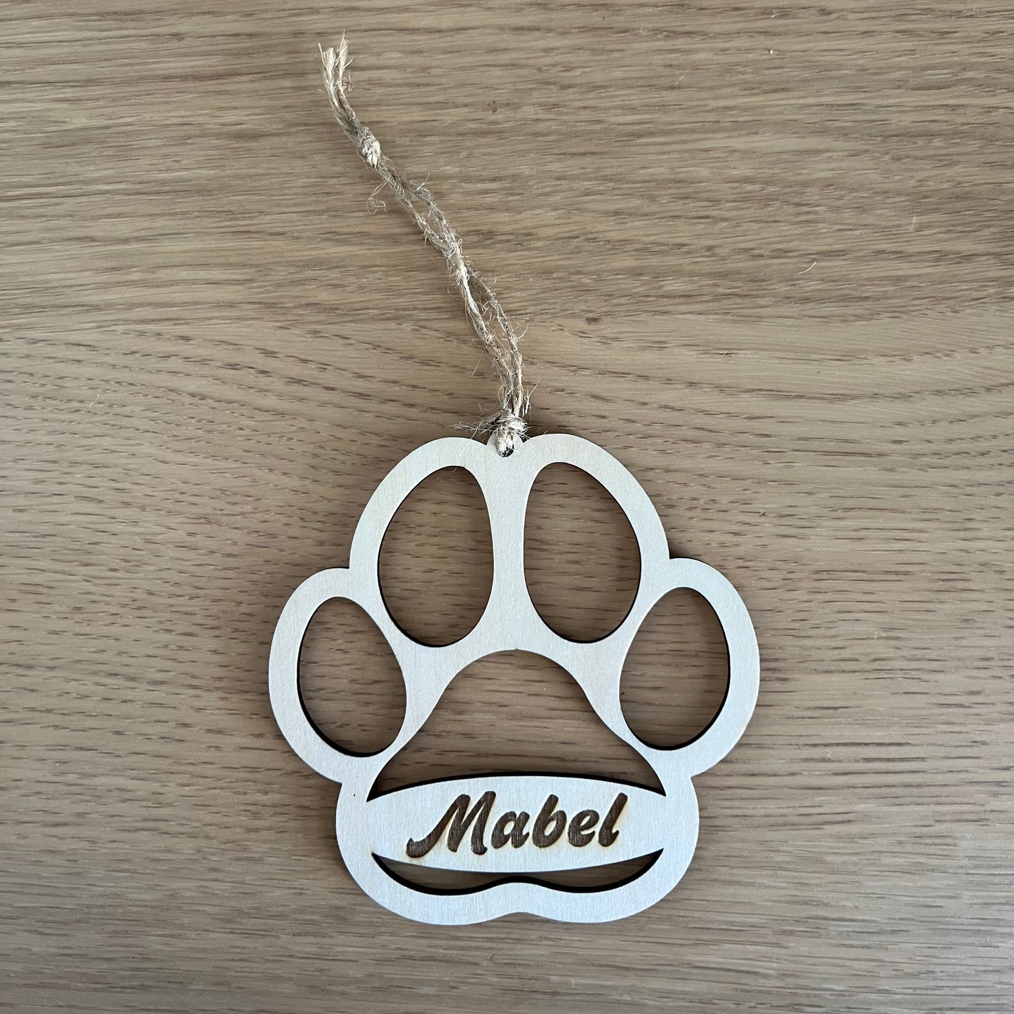 A hand hangs a personalized Dog Paw ornament by Wet Nose Woodworks with the name Mabel on a green Christmas tree, adding a unique touch to your holiday decor.