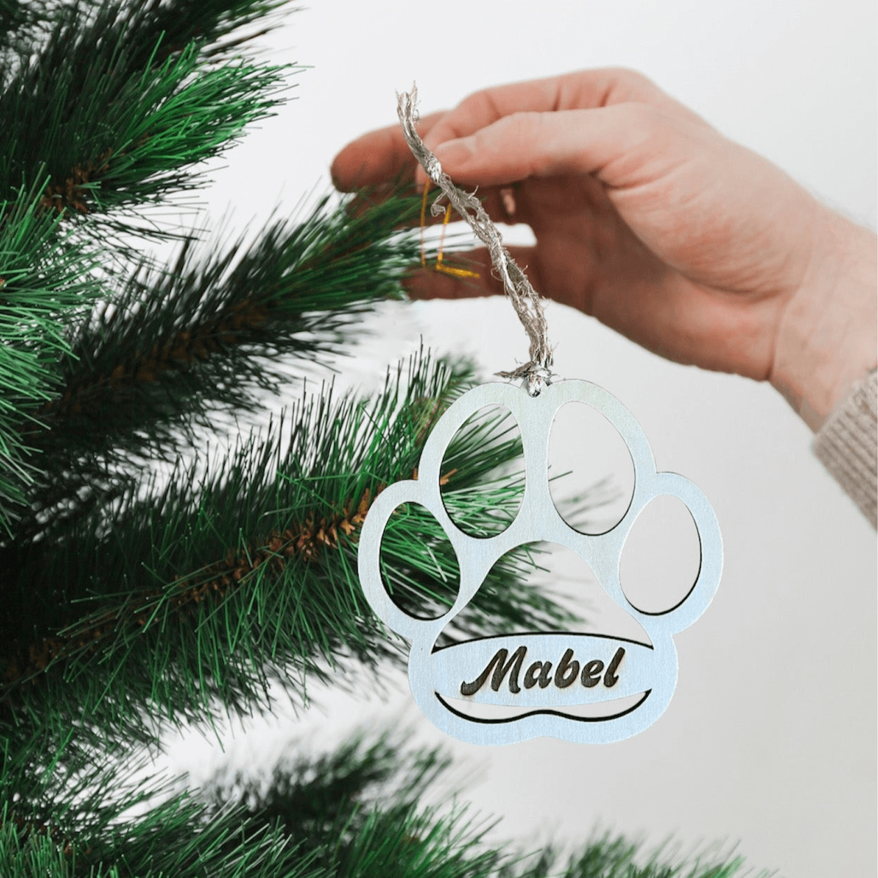 A hand hangs a personalized Dog Paw ornament by Wet Nose Woodworks with the name Mabel on a green Christmas tree, adding a unique touch to your holiday decor.
