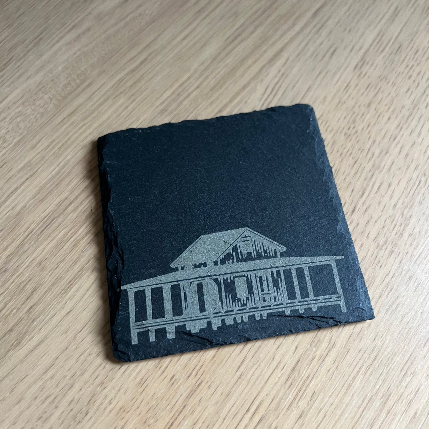 The Grass Island Coaster by Wet Nose Woodworks, featuring a white etched image of the iconic Guilford, CT shack on black slate, rests on wood