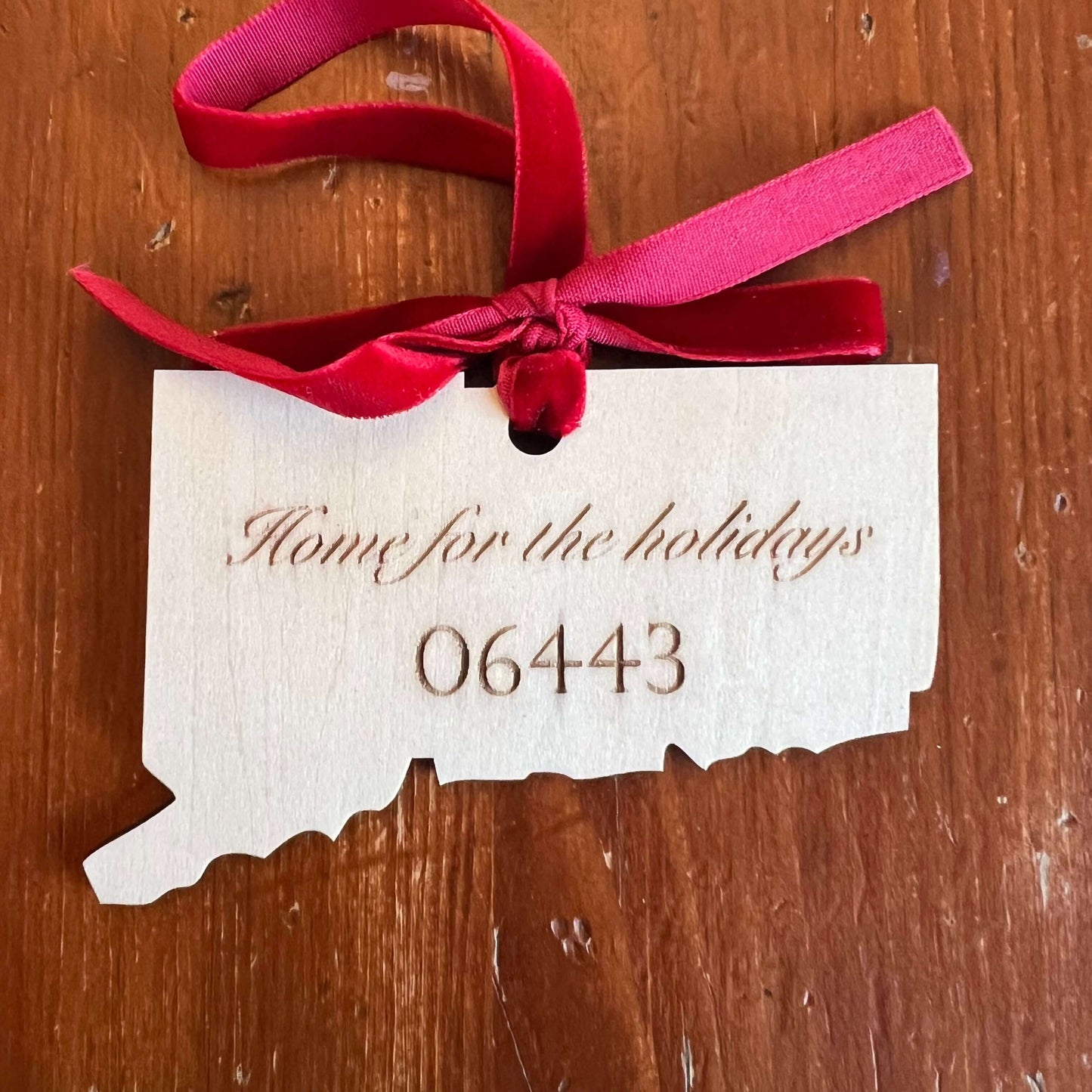 The Connecticut State Ornament with Personalization by Wet Nose Woodworks is a handcrafted wooden keepsake engraved with Home for the holidays and zip code 06437, tied with a gold ribbon to showcase Connecticut pride.