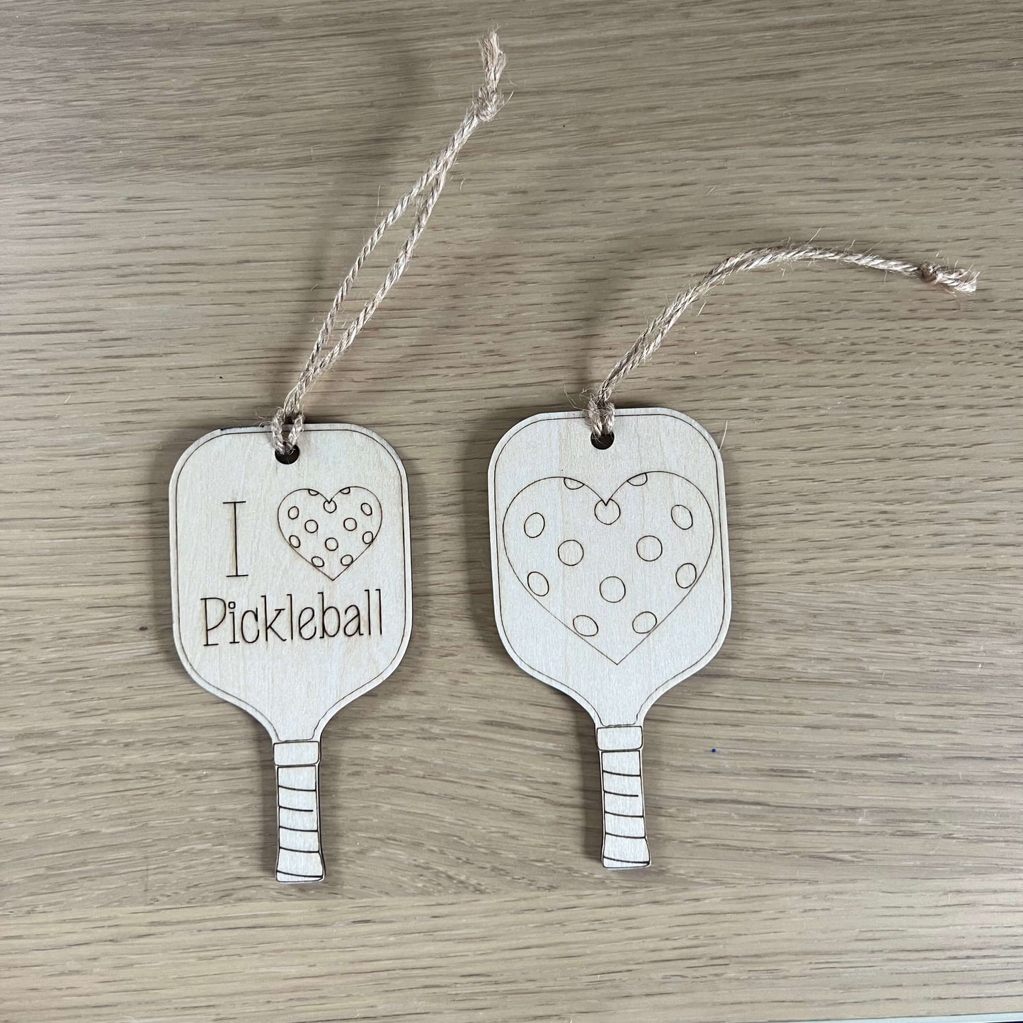 The I Heart Pickleball Ornament by Wet Nose Woodworks features two wooden pickleball paddle tags with strings—one saying I ♥ Pickleball and the other with a polka dot heart. A unique, charming gift for any pickleball enthusiast.