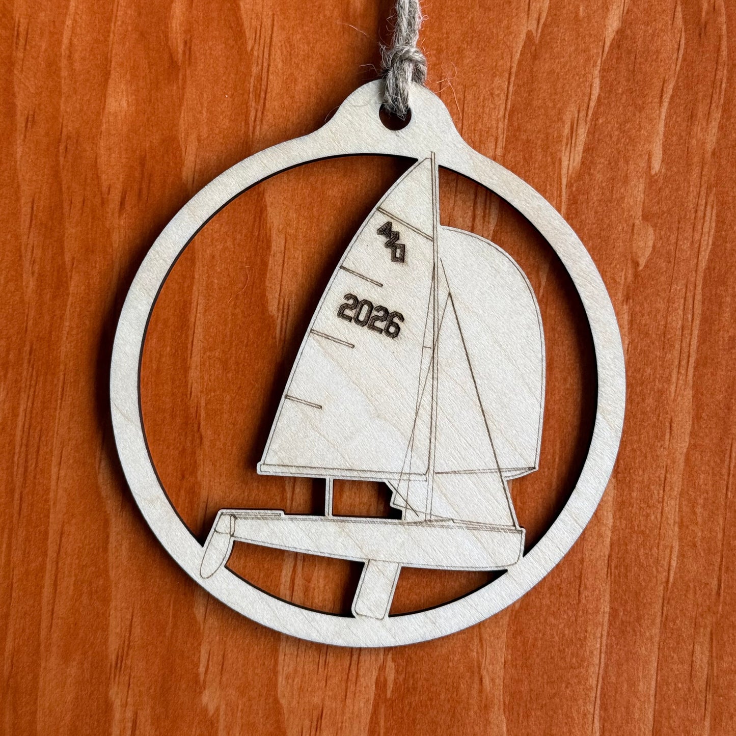 A laser-cut wooden ornament shaped like a sailboat inside a circular frame, hanging from a twine string against a warm wood-grain background. The sail features the text '2026'.