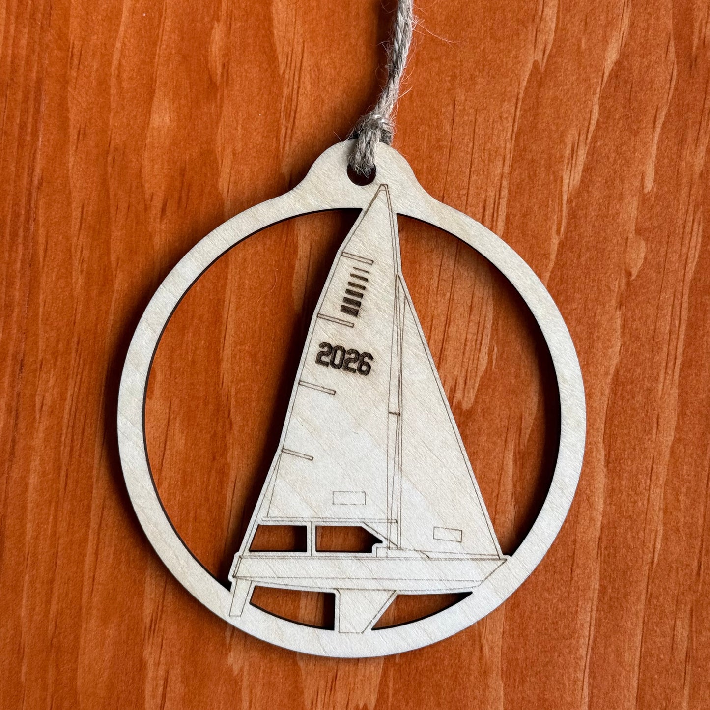 A wooden sailboat ornament inside a circular frame with a hanging twine string, displayed on a warm brown wood-grain background. The sailboat features the engraved year '2026'.