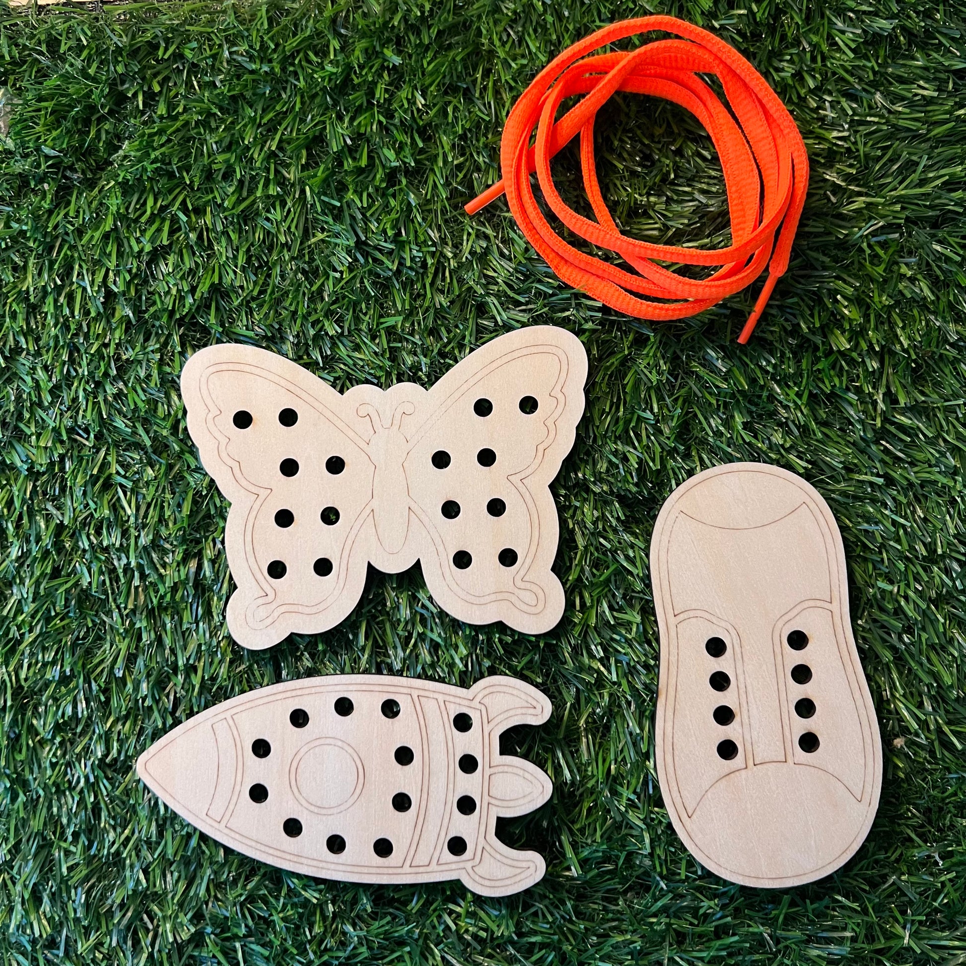 The Wet Nose Woodworks Lacing Toy Small Set includes three wooden boards—a butterfly, rocket, and shoe—each with holes for threading, shown on green grass beside an orange shoelace to boost fine motor skills.