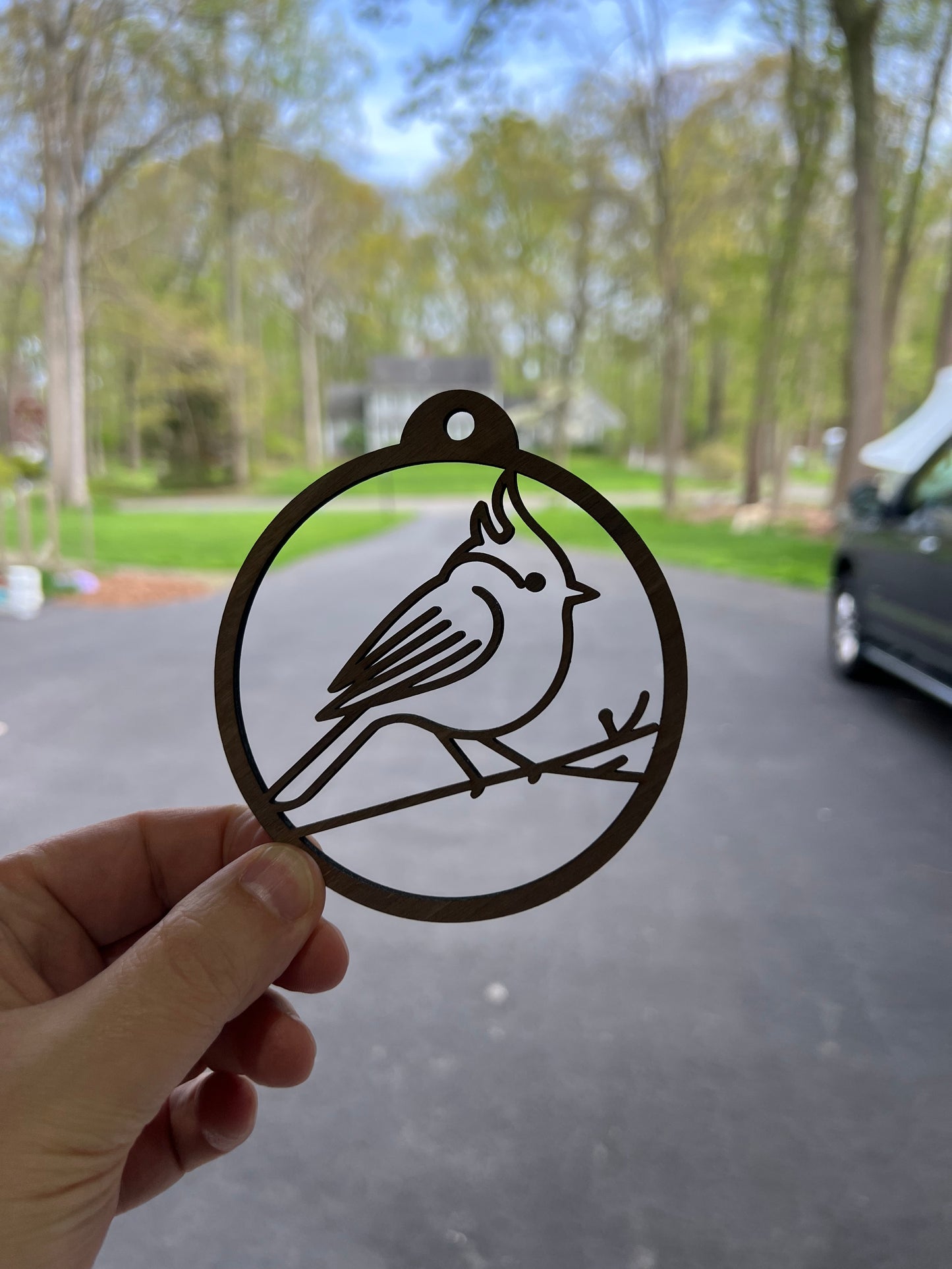 The Wet Nose Woodworks Tufted Titmouse ornament, a black metal bird perched on a branch, hangs on the window for rustic charm, with a blurred green yard and trees outside.