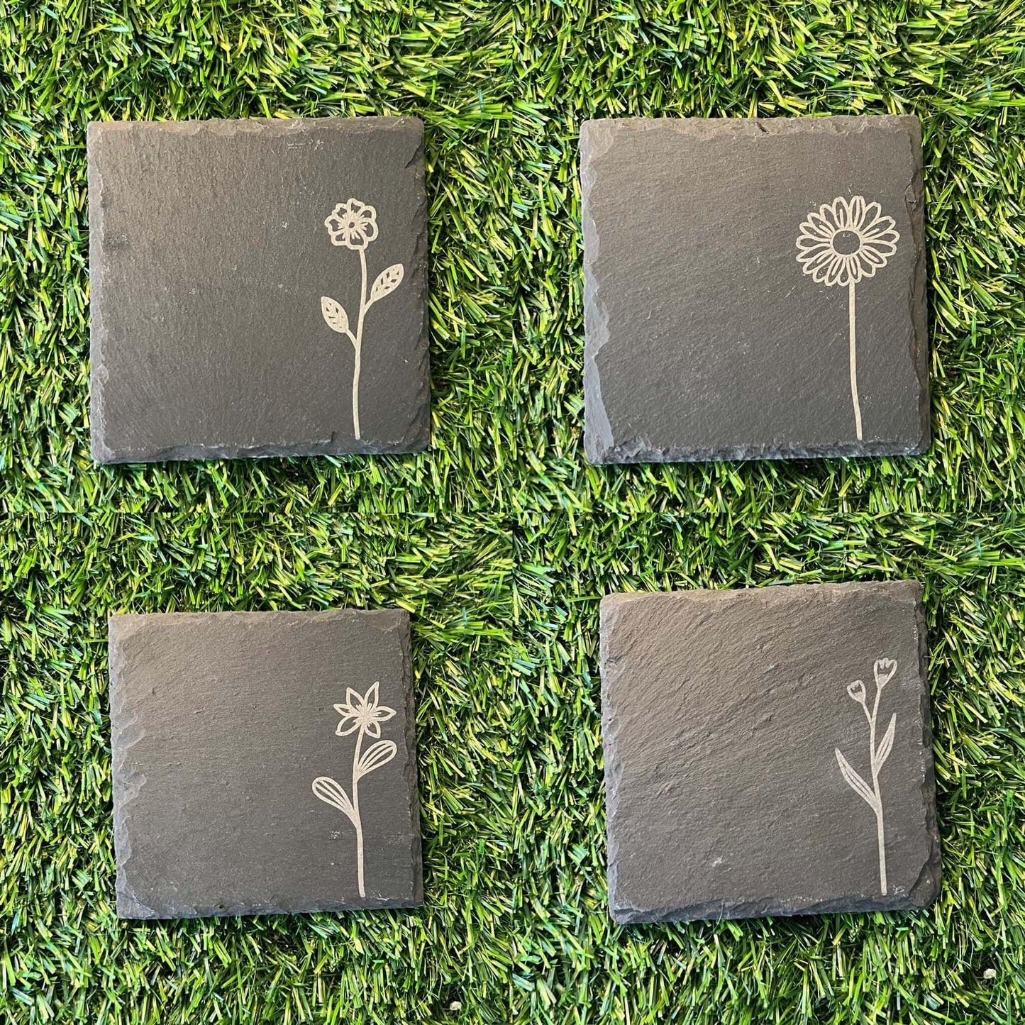 Four Wildflower Coasters by Wet Nose Woodworks—three with engraved wildflowers and one plain—are arranged in a square on green artificial grass. Each black slate coaster highlights natural beauty and craftsmanship.