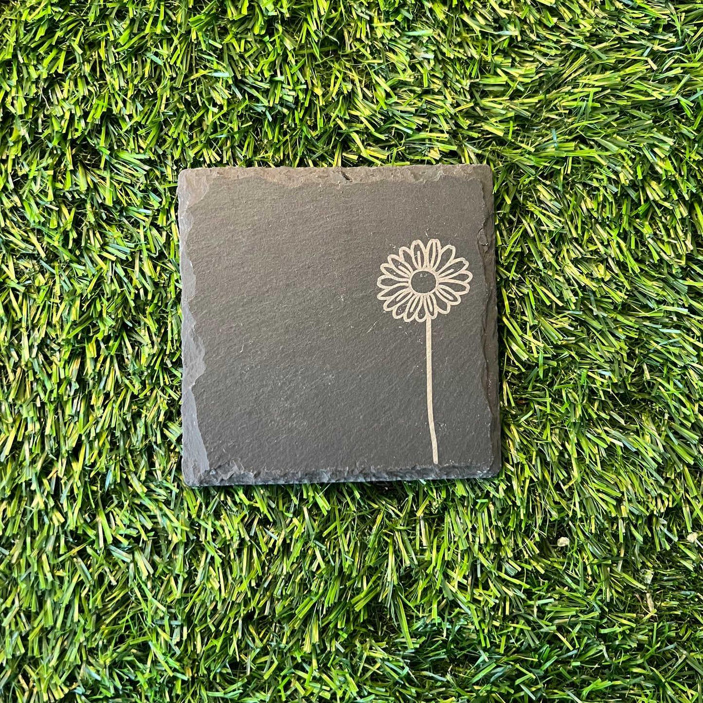 Four Wildflower Coasters by Wet Nose Woodworks—three with engraved wildflowers and one plain—are arranged in a square on green artificial grass. Each black slate coaster highlights natural beauty and craftsmanship.