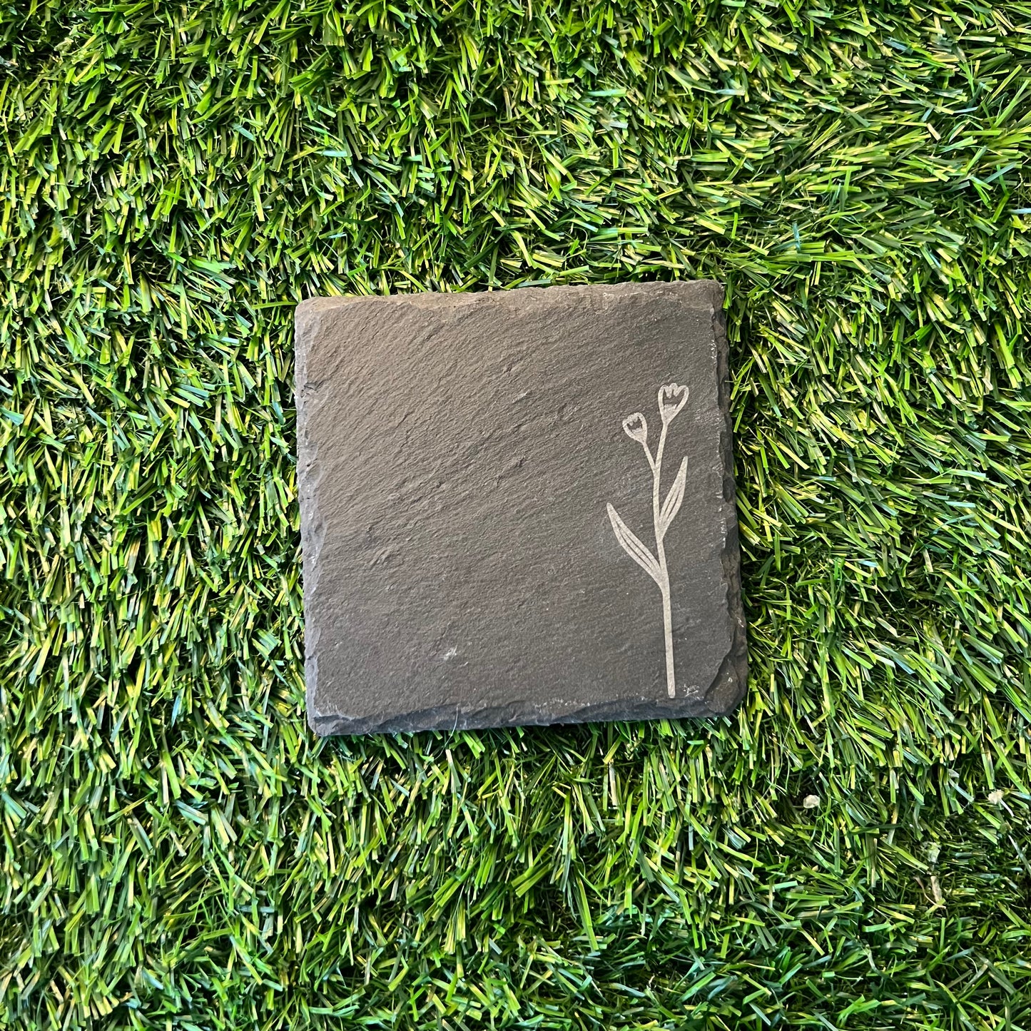 Four Wildflower Coasters by Wet Nose Woodworks—three with engraved wildflowers and one plain—are arranged in a square on green artificial grass. Each black slate coaster highlights natural beauty and craftsmanship.