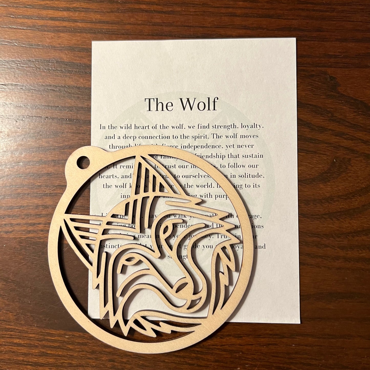 The Wolf Ornament by Wet Nose Woodworks, ideal for rustic home decor, sits atop a printed sheet titled “The Wolf” that displays wolf-themed text—all displayed on a wooden table.