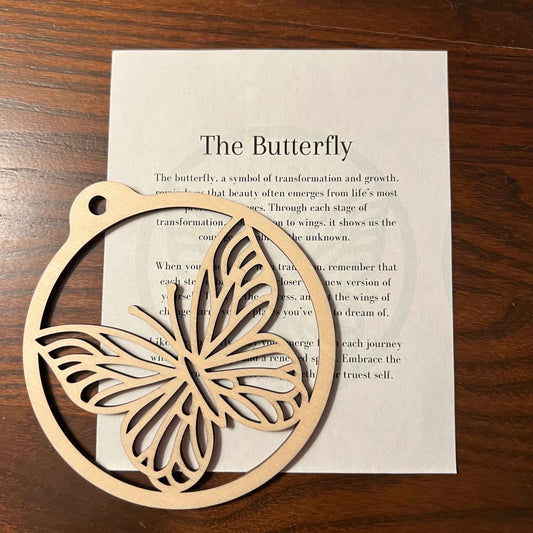 The Butterfly Ornament by Wet Nose Woodworks sits atop a “The Butterfly” card with transformation text, offering a charming rustic accent for your home on a dark wood surface.
