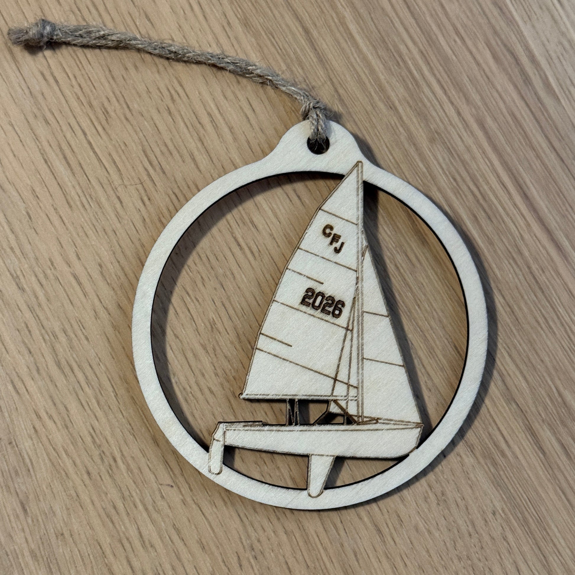 A circular wooden ornament featuring a laser-cut sailboat design with the engraved text 'CFJ' and '2026', hanging from a rustic twine string on a light wood grain surface.