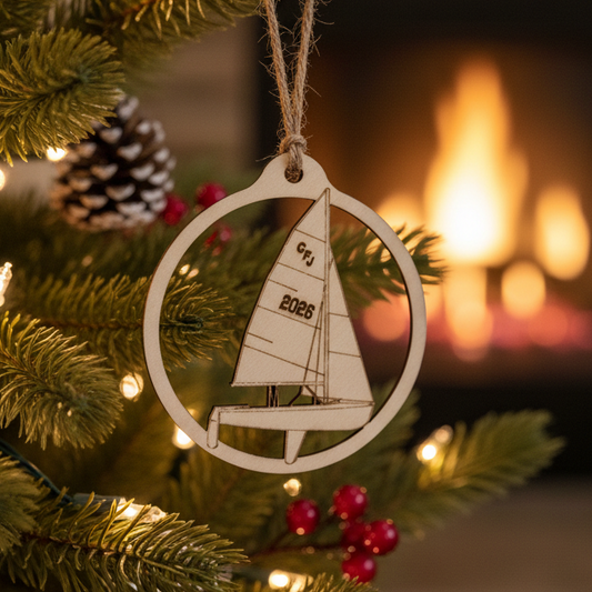 A wooden laser-cut Christmas ornament shaped like a sailboat inside a circle, hanging on a decorated Christmas tree with warm lights and a blurred fireplace in the background.