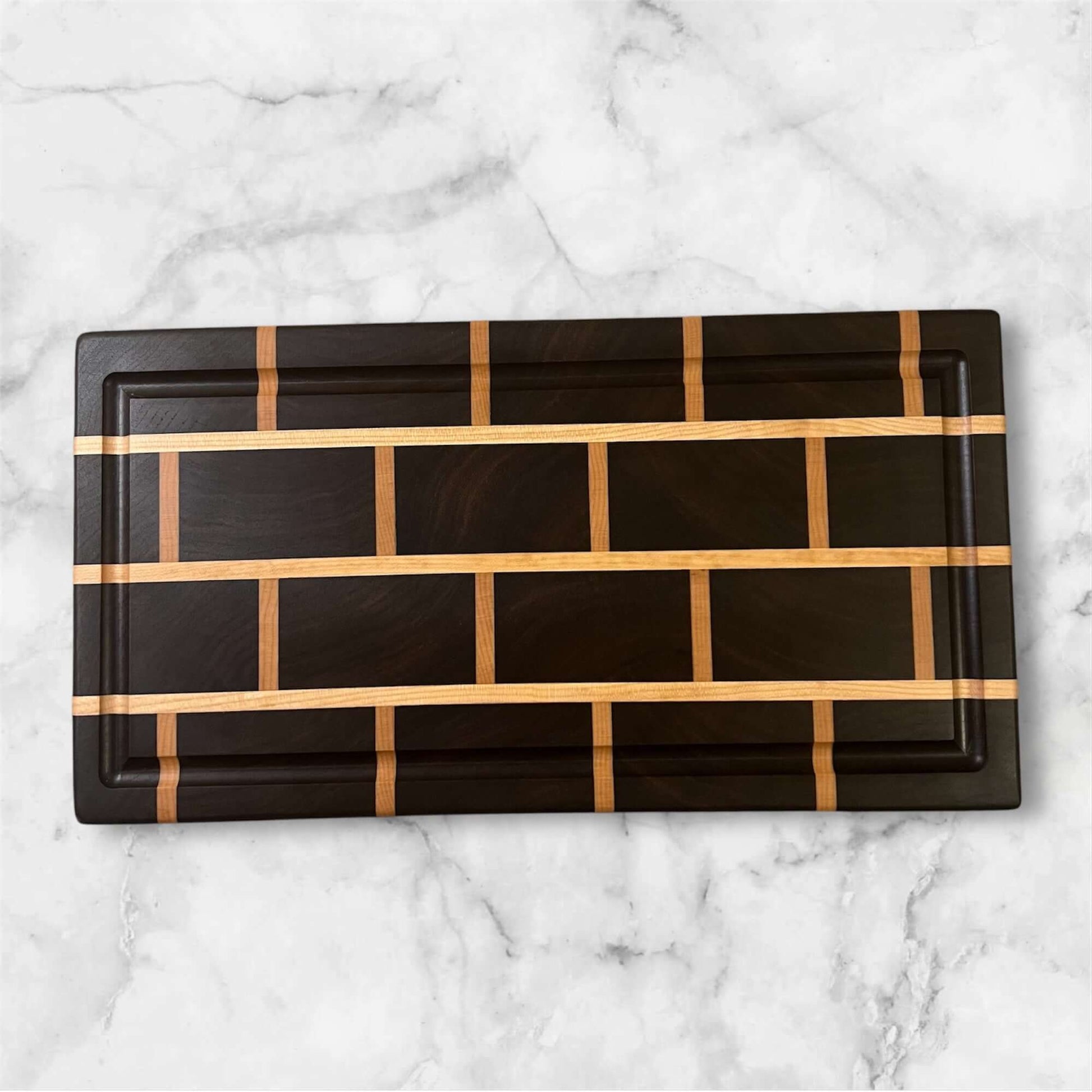 The Artisan Board #8 by Wet Nose Woodworks is a rectangular walnut and maple cutting board featuring a brick wall pattern of alternating dark and light wood strips, displayed on a white marble surface.
