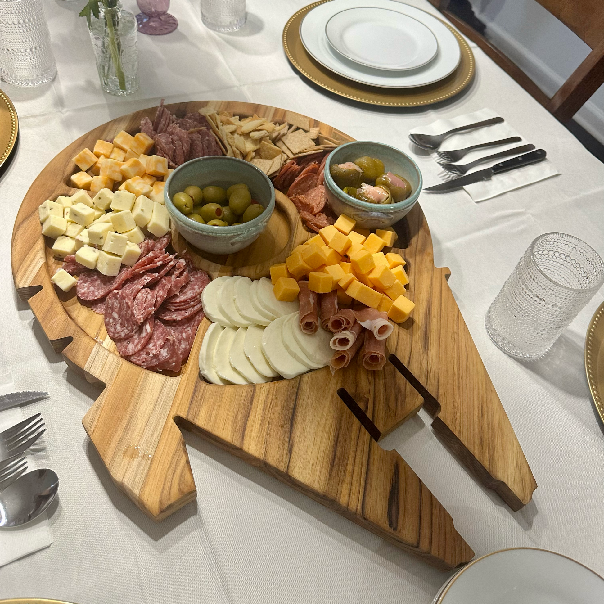 A large wooden charcuterie board shaped like a spaceship, filled with sliced cheeses, cured meats, crackers, olives, and rolled prosciutto, set on a dining table surrounded by plates, cutlery, and glasses.