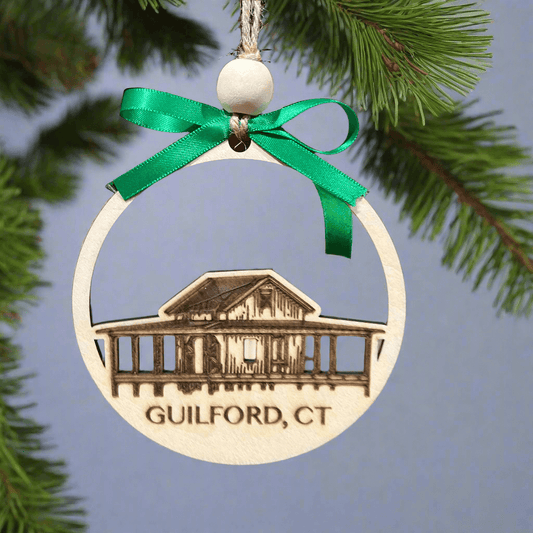 The Grass Island Shack Ornament by Wet Nose Woodworks features a round wooden design with a laser-etched image of the iconic shack and Guilford, CT. It hangs from a tree branch with a green ribbon and white bead.