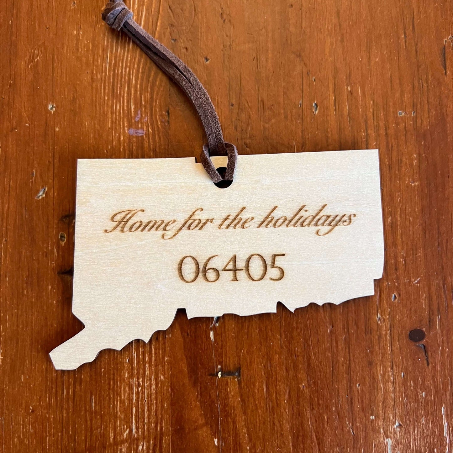 The Connecticut State Ornament with Personalization by Wet Nose Woodworks is a handcrafted wooden keepsake engraved with Home for the holidays and zip code 06437, tied with a gold ribbon to showcase Connecticut pride.