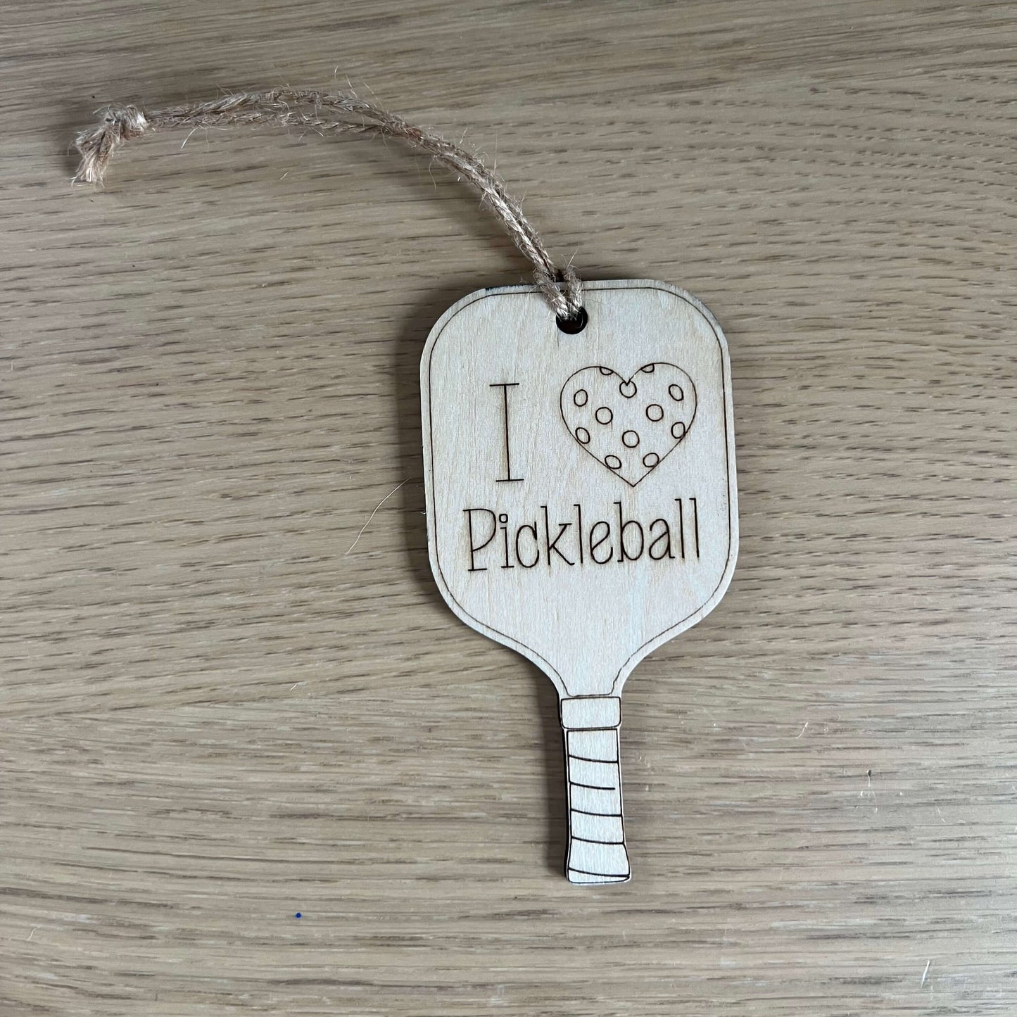 The I Heart Pickleball Ornament by Wet Nose Woodworks features two wooden pickleball paddle tags with strings—one saying I ♥ Pickleball and the other with a polka dot heart. A unique, charming gift for any pickleball enthusiast.
