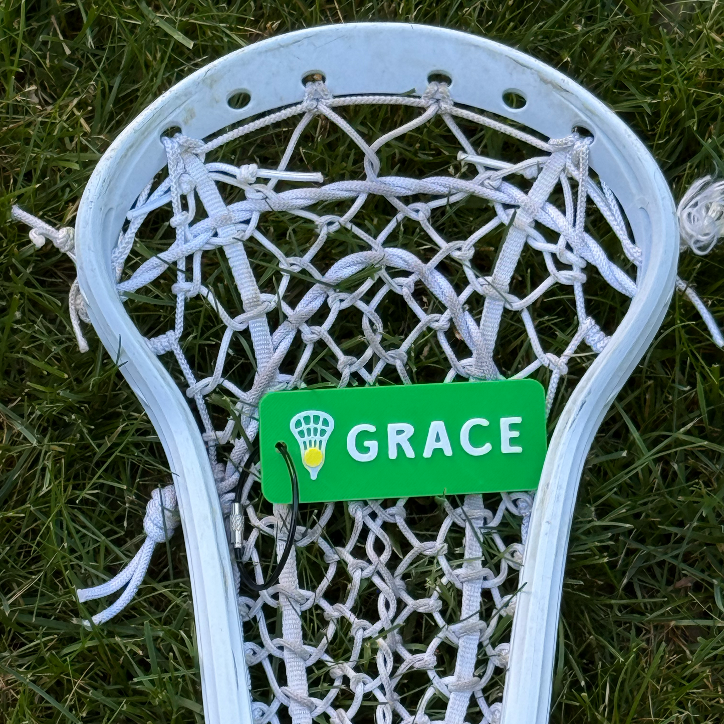 Personalized green lacrosse bag tag attached to a lacrosse head on grass