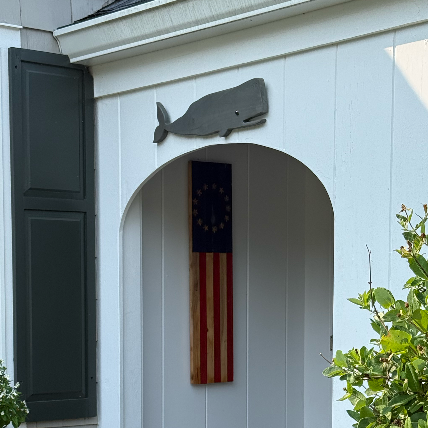 A vertical shot of a white house exterior featuring a decorative American flag with a gold border mounted on the arched entryway, flanked by a gray whale silhouette and green shutters.