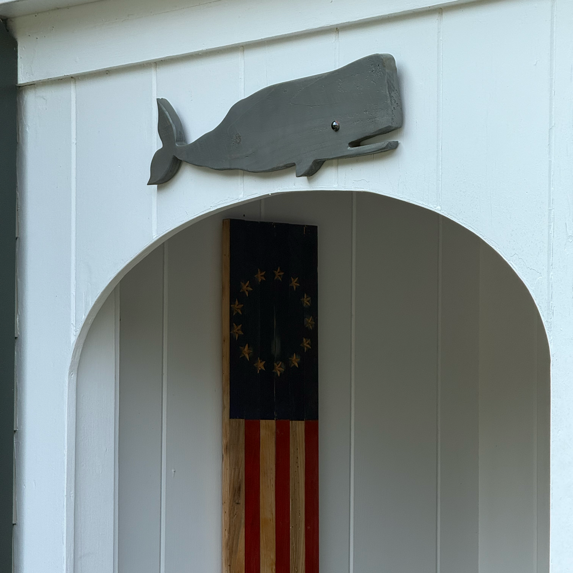 A rustic wooden wall plaque featuring a stylized American flag design with a navy blue canton and red and brown stripes, mounted on a white paneled wall next to a gray whale silhouette decoration.
