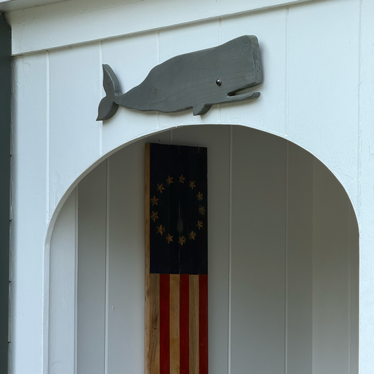 A rustic wooden wall plaque featuring a stylized American flag design with a navy blue canton and red and brown stripes, mounted on a white paneled wall next to a gray whale silhouette decoration.