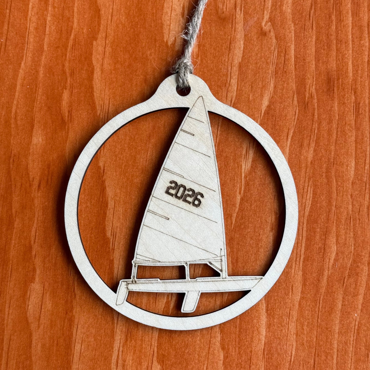A silver metal ornament shaped like a sailboat inside a circular frame, with a gray string attached and the year '2026' engraved on the sail, displayed on a warm brown wood background.