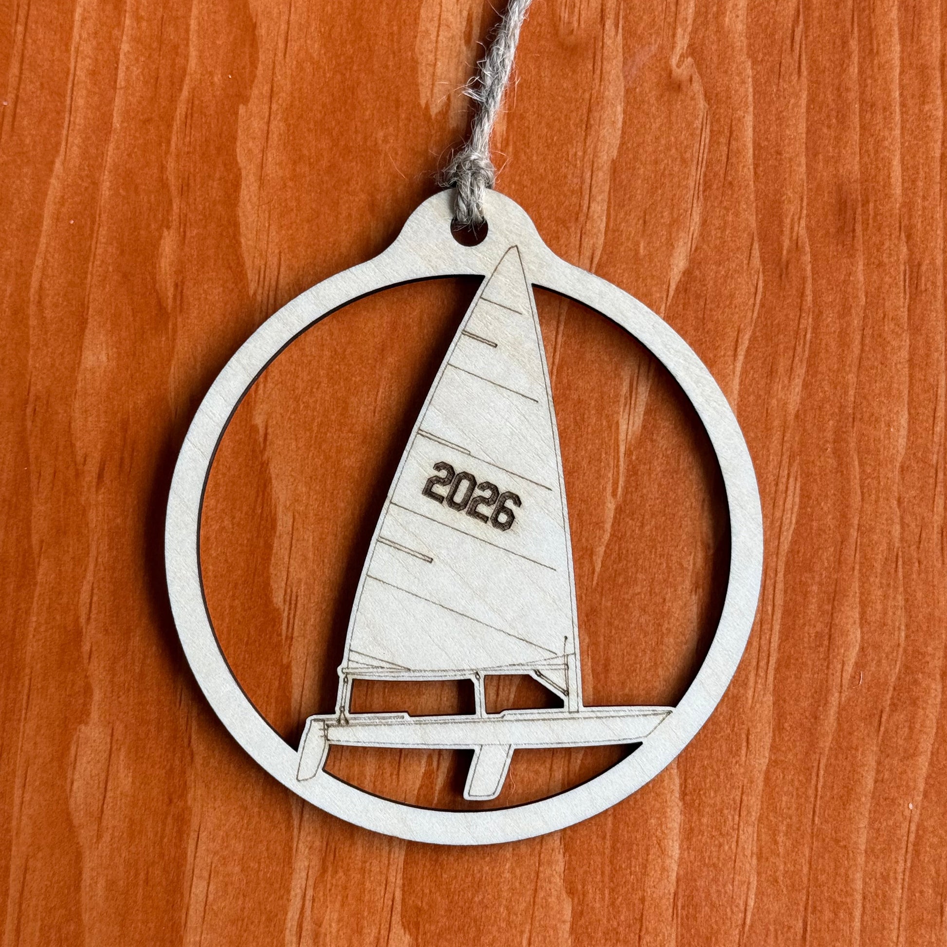 A silver metal ornament shaped like a sailboat inside a circular frame, with a gray string attached and the year '2026' engraved on the sail, displayed on a warm brown wood background.