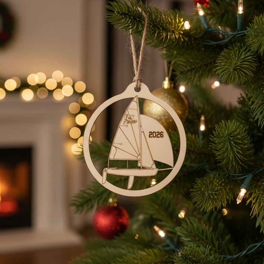 A wood ornament shaped like a sailboat inside a circular frame, hanging from a gray string against a warm brown wooden background. The sail features the word 'RS Feva' and the year '2026' engraved.