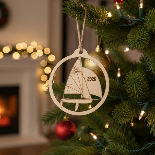 A wood ornament shaped like a sailboat inside a circular frame, hanging from a gray string against a warm brown wooden background. The sail features the word 'RS Feva' and the year '2026' engraved.