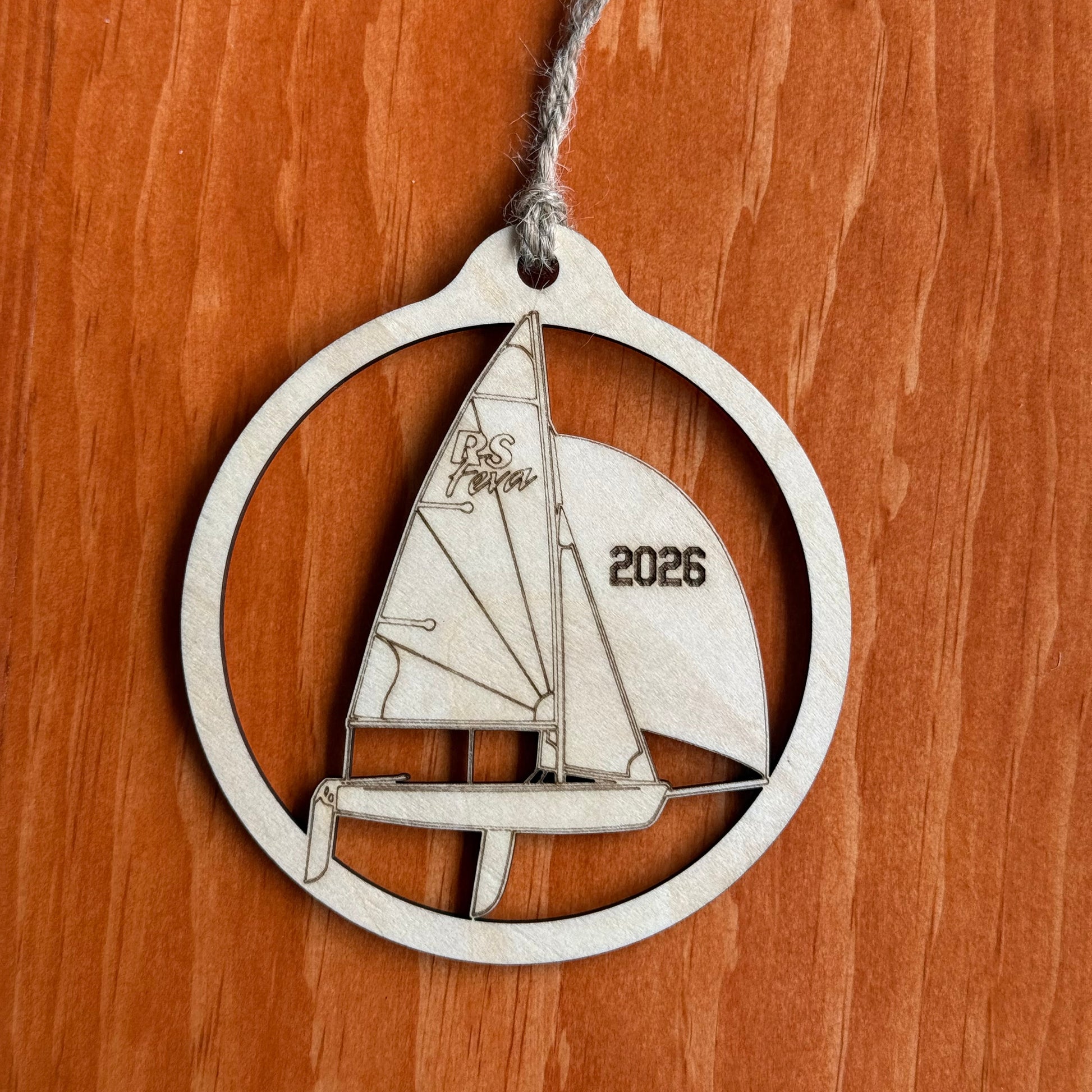 A wooden ornament shaped like a sailboat inside a circular frame, hanging from a gray string against a warm brown wooden background. The sail features the word 'RS Feva' and the year '2026' engraved.