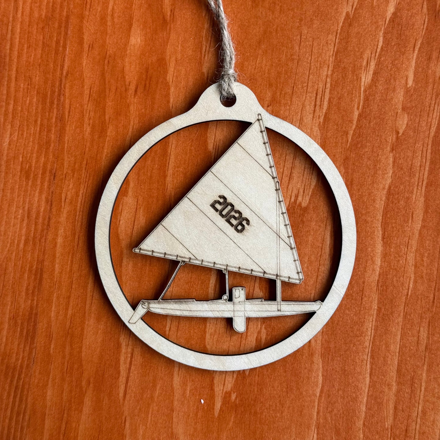 A wooden ornament shaped like a sailboat inside a circular frame, with a hanging twine string and the year '2026' printed on the sail, shown on a warm brown wood surface.