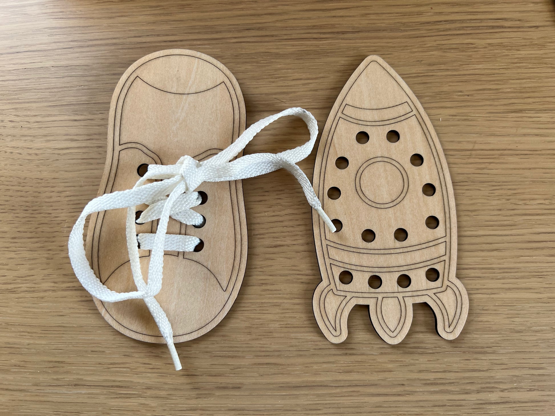 Two Wet Nose Woodworks Lacing Toys on a wooden surface: one shoe-shaped with a white lace for knot tying and fine motor skills, and one rocket-shaped with lacing holes but no string included.