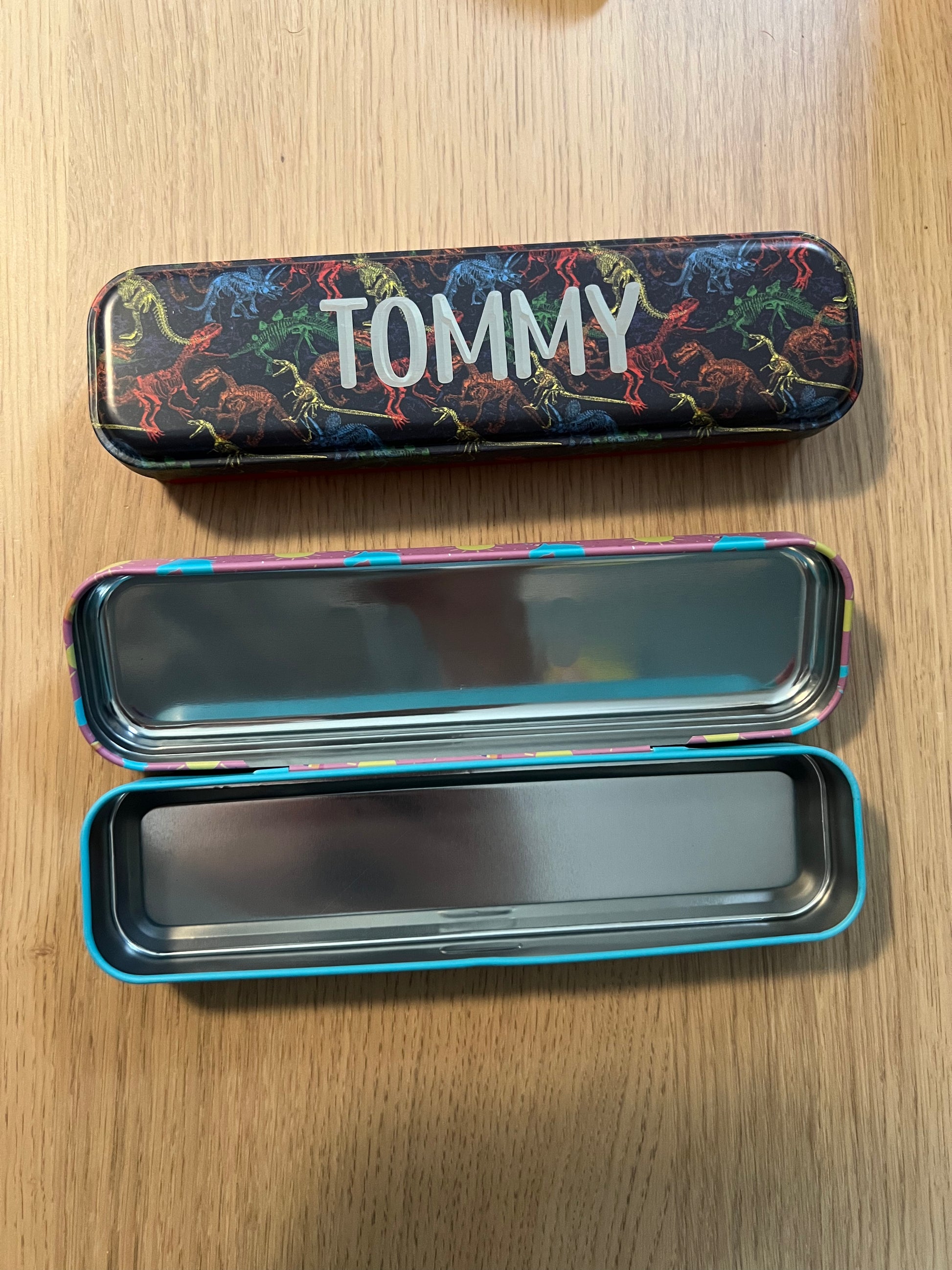 Wet Nose Woodworks Metal Pencil Boxes come in four colorful designs—Kaylea (animal print), Celia (striped), Grace (rainbows & crowns), and Tommy (dinosaurs)—with snap closures, keeping desks stylish and organized.