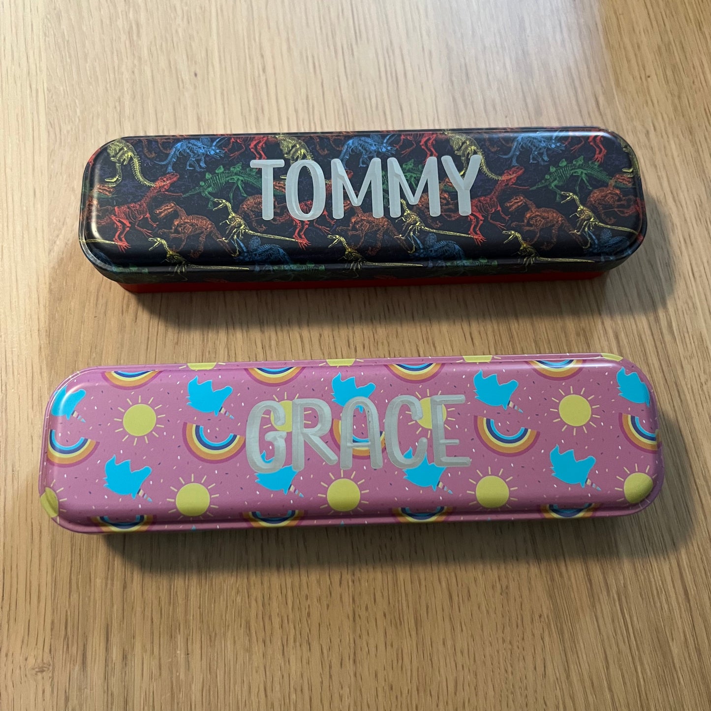 Wet Nose Woodworks Metal Pencil Boxes come in four colorful designs—Kaylea (animal print), Celia (striped), Grace (rainbows & crowns), and Tommy (dinosaurs)—with snap closures, keeping desks stylish and organized.
