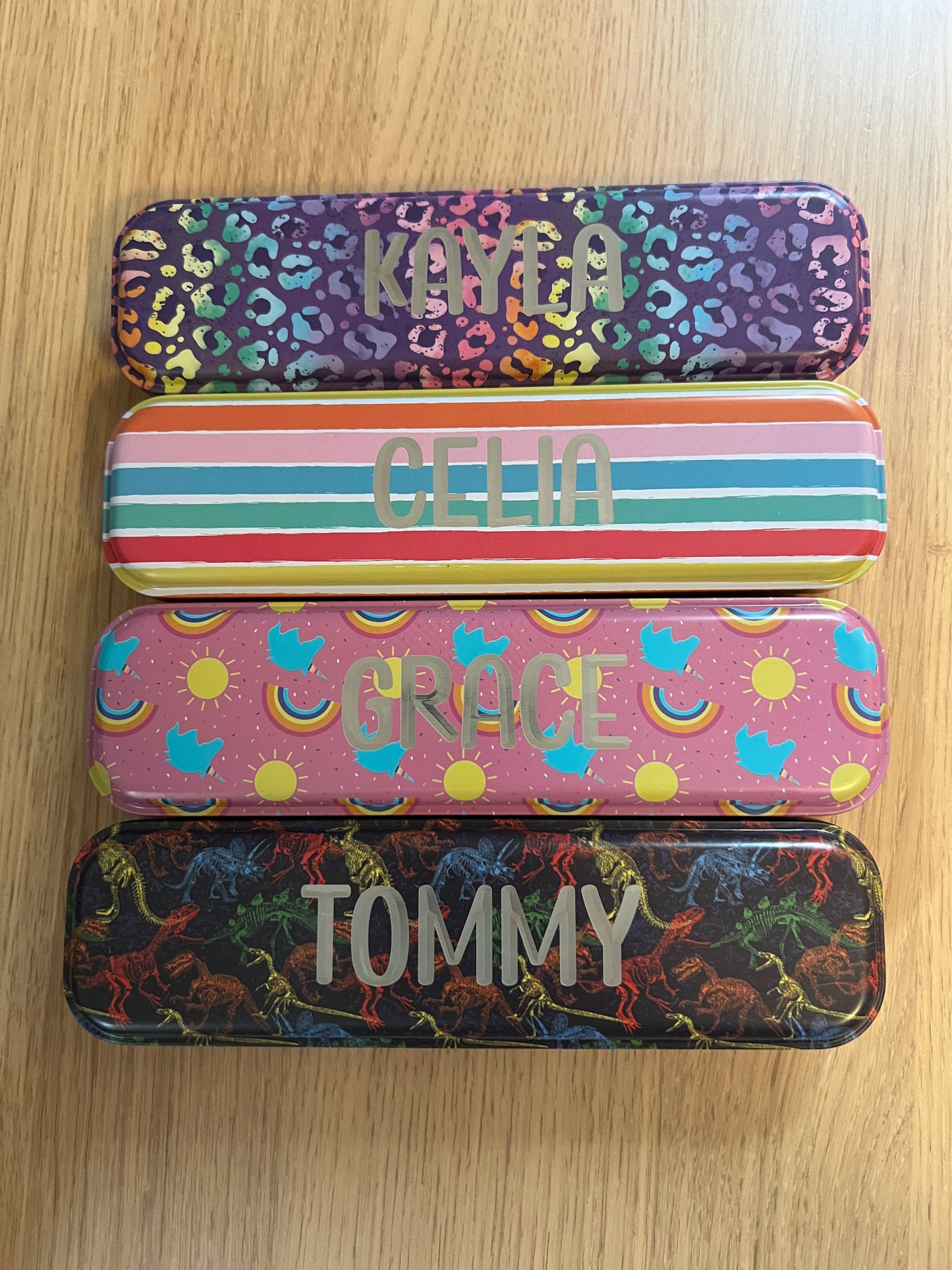 Wet Nose Woodworks Metal Pencil Boxes come in four colorful designs—Kaylea (animal print), Celia (striped), Grace (rainbows & crowns), and Tommy (dinosaurs)—with snap closures, keeping desks stylish and organized.