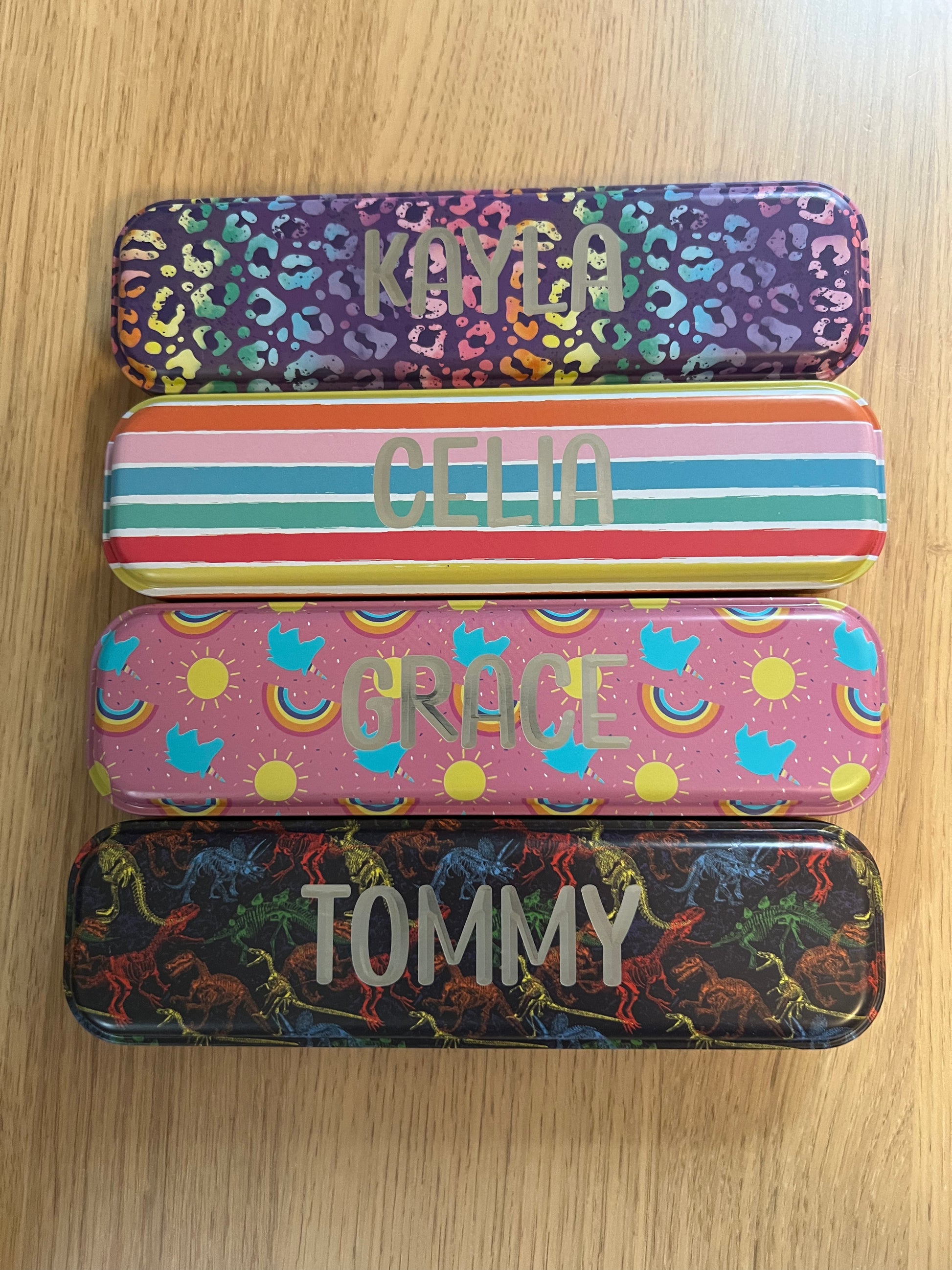 Wet Nose Woodworks Metal Pencil Boxes come in four colorful designs—Kaylea (animal print), Celia (striped), Grace (rainbows & crowns), and Tommy (dinosaurs)—with snap closures, keeping desks stylish and organized.