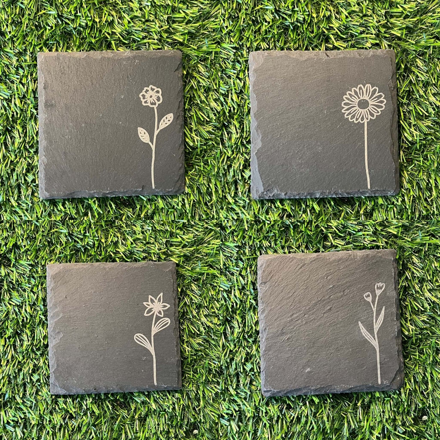 Four Wildflower Coasters by Wet Nose Woodworks—three with engraved wildflowers and one plain—are arranged in a square on green artificial grass. Each black slate coaster highlights natural beauty and craftsmanship.