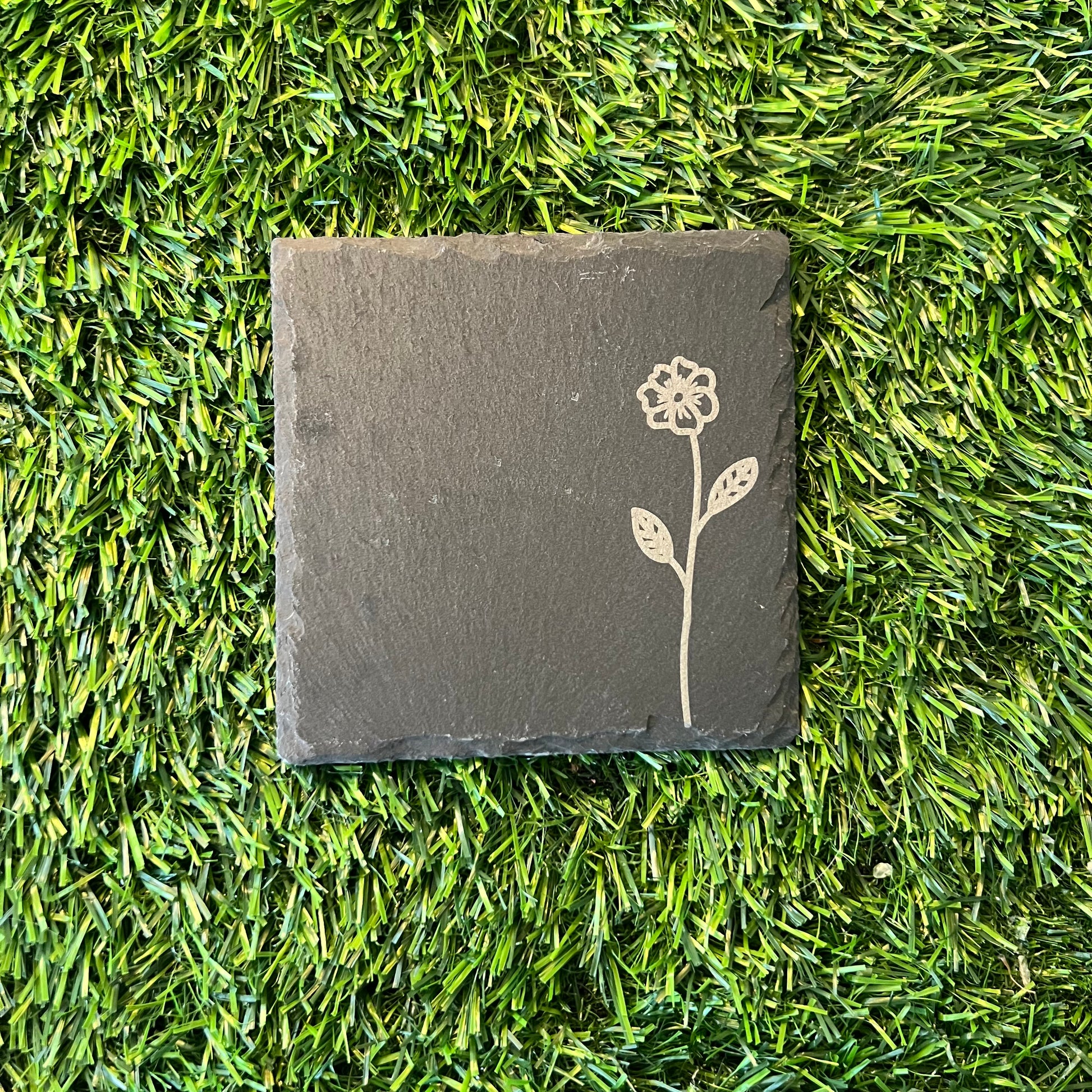 Four Wildflower Coasters by Wet Nose Woodworks—three with engraved wildflowers and one plain—are arranged in a square on green artificial grass. Each black slate coaster highlights natural beauty and craftsmanship.