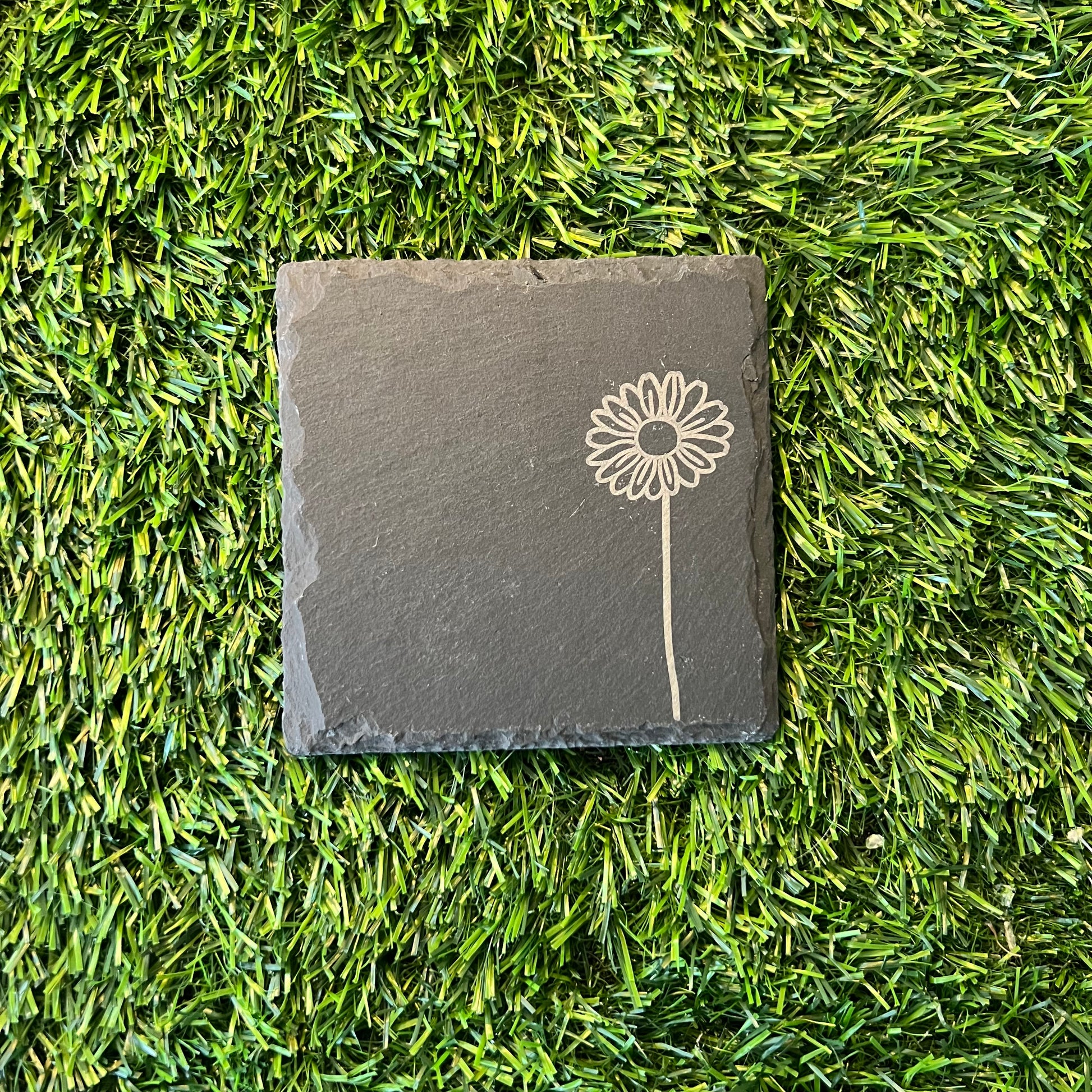 Four Wildflower Coasters by Wet Nose Woodworks—three with engraved wildflowers and one plain—are arranged in a square on green artificial grass. Each black slate coaster highlights natural beauty and craftsmanship.