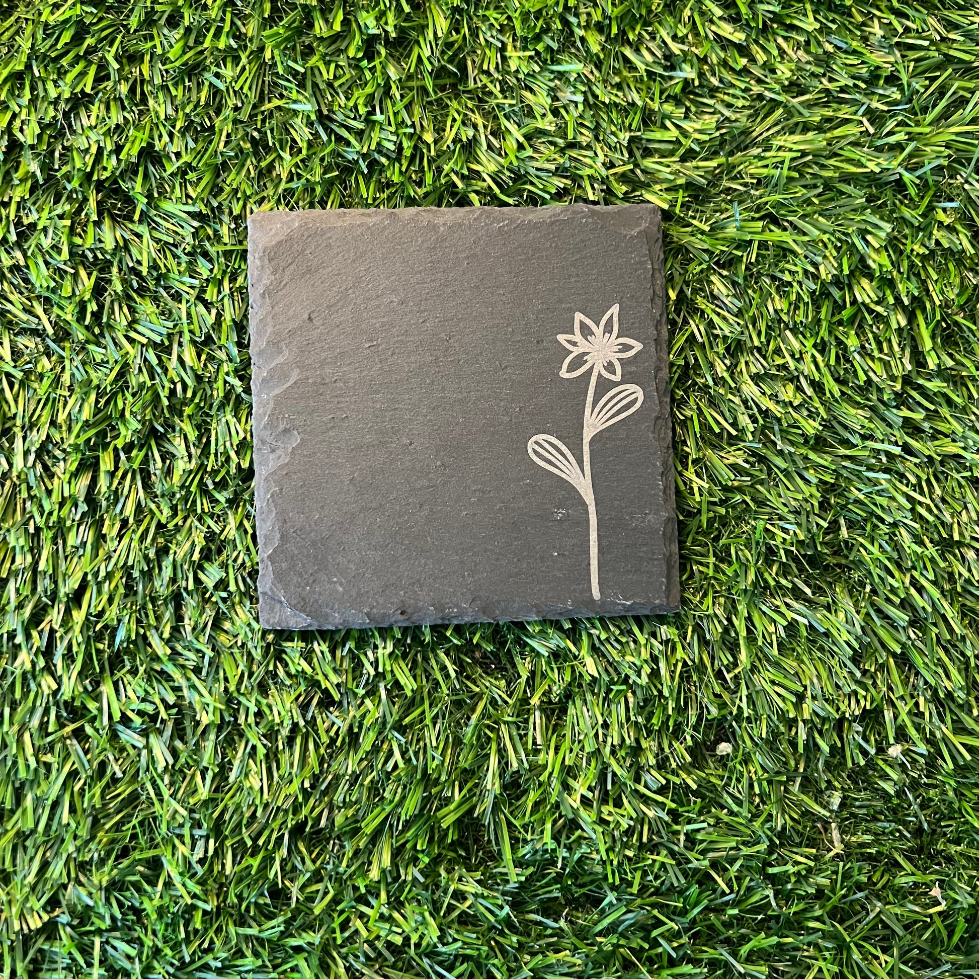 Four Wildflower Coasters by Wet Nose Woodworks—three with engraved wildflowers and one plain—are arranged in a square on green artificial grass. Each black slate coaster highlights natural beauty and craftsmanship.
