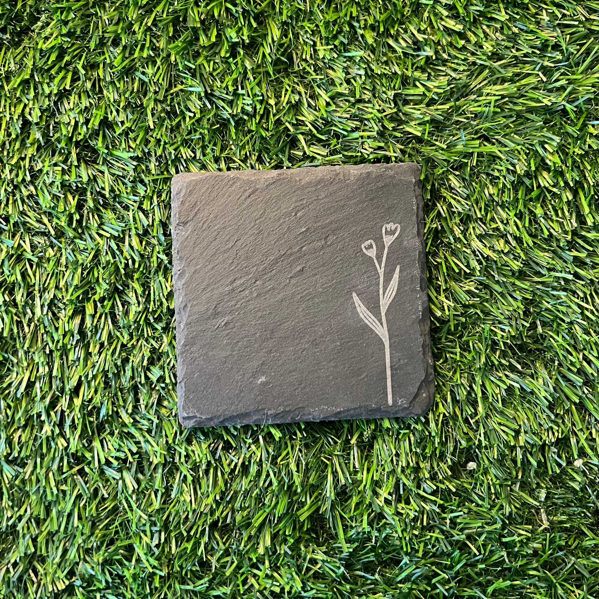 Four Wildflower Coasters by Wet Nose Woodworks—three with engraved wildflowers and one plain—are arranged in a square on green artificial grass. Each black slate coaster highlights natural beauty and craftsmanship.